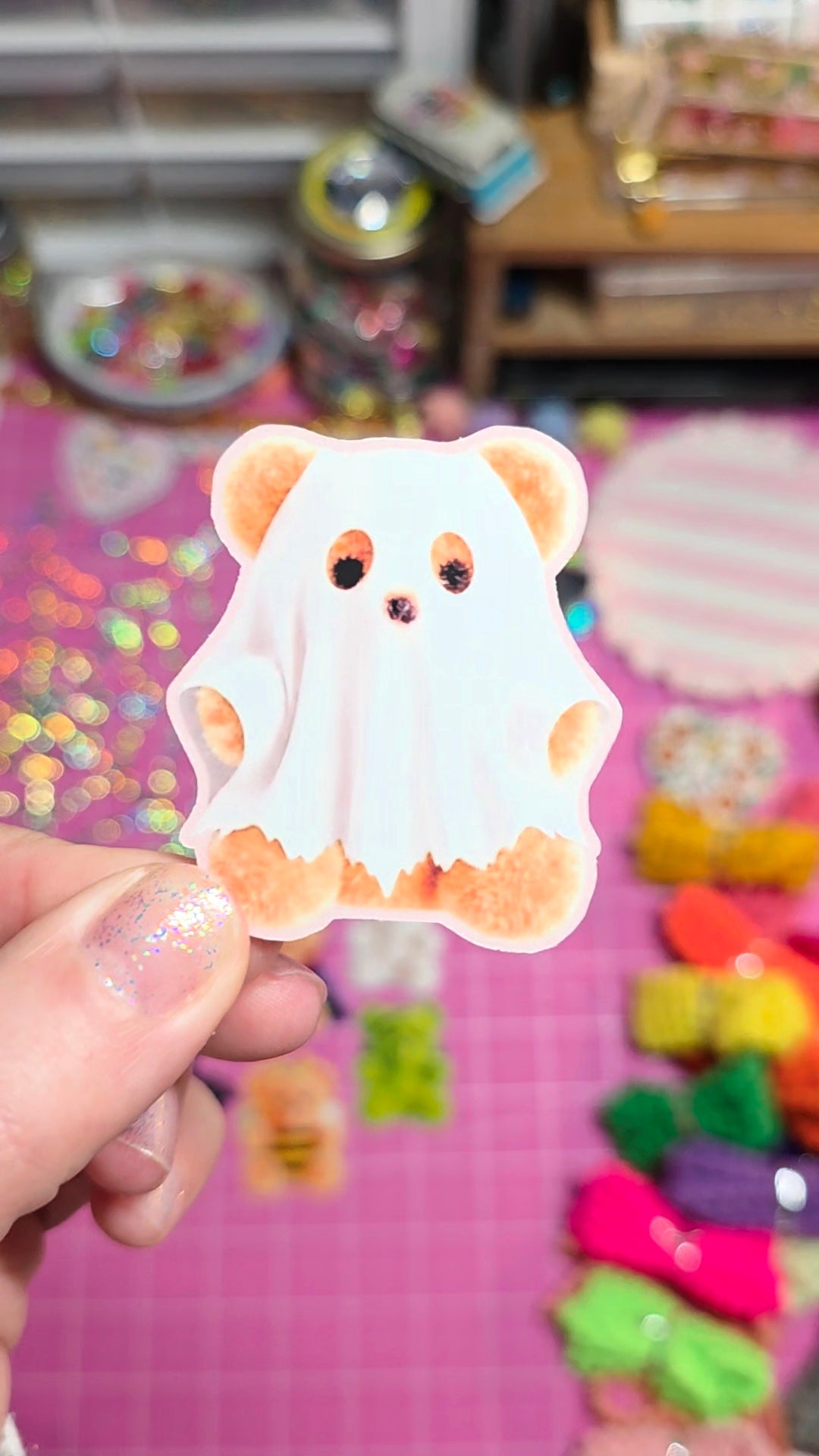 A hand holding a cute ghost-shaped sticker or decal featuring a cartoon bear face with orange eyes and paws, set against a colorful, blurred craft background.