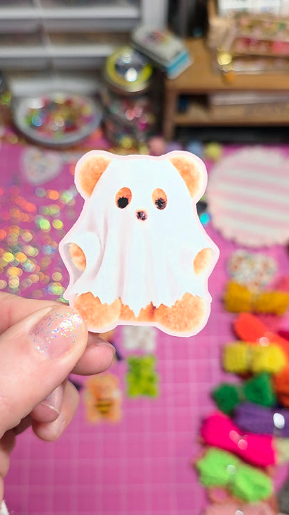 A hand holding a cute ghost-shaped sticker or decal featuring a cartoon bear face with orange eyes and paws, set against a colorful, blurred craft background.