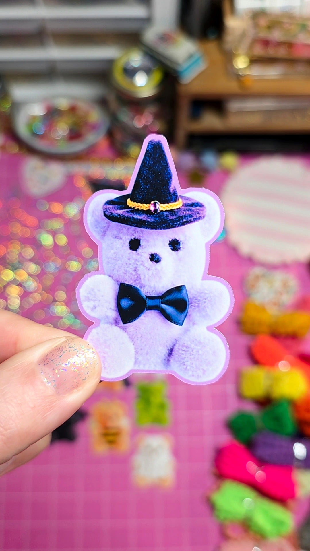 A hand holds a small fuzzy lavender teddy bear charm wearing a glittery black witch hat and dark blue bow tie, against a colorful pink background with craft supplies.