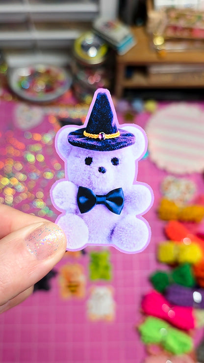 A hand holds a small fuzzy lavender teddy bear charm wearing a glittery black witch hat and dark blue bow tie, against a colorful pink background with craft supplies.