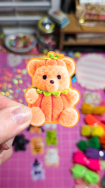 A hand with glittery nail polish holds a small plush teddy bear shaped like a pumpkin, featuring orange faux fur, a green leafy stem, and a green bow tie, against a colorful, blurred background of craft supplies.
