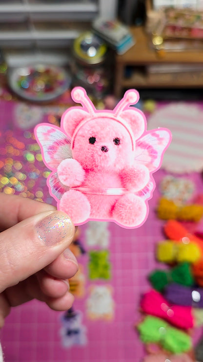 A hand holds a small pink fuzzy teddy bear accessory with butterfly wings and antennae against a colorful, blurred background.