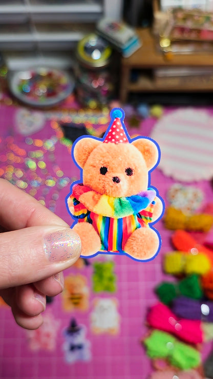 A hand holds a small plush teddy bear accessory with a colorful rainbow outfit and party hat, set against a busy pink craft workspace background.