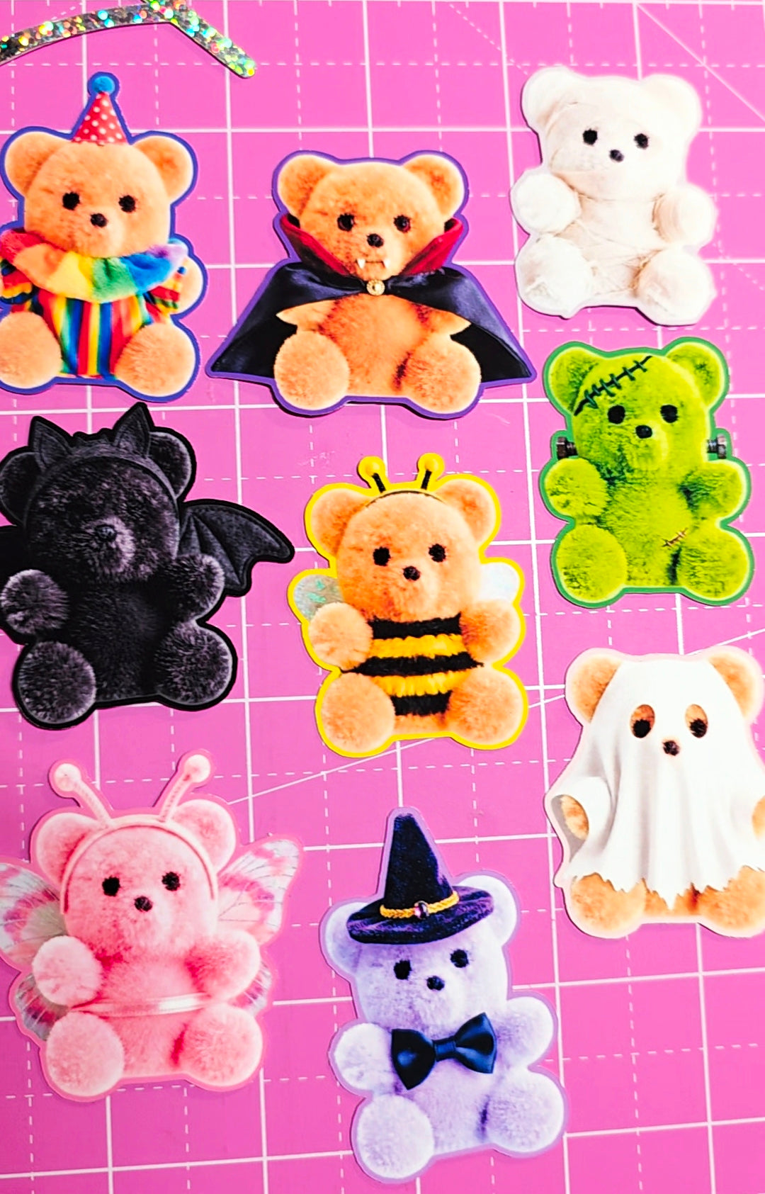 A collection of plush teddy bear appliques in various Halloween and character designs, arranged on a pink cutting mat with grid lines.