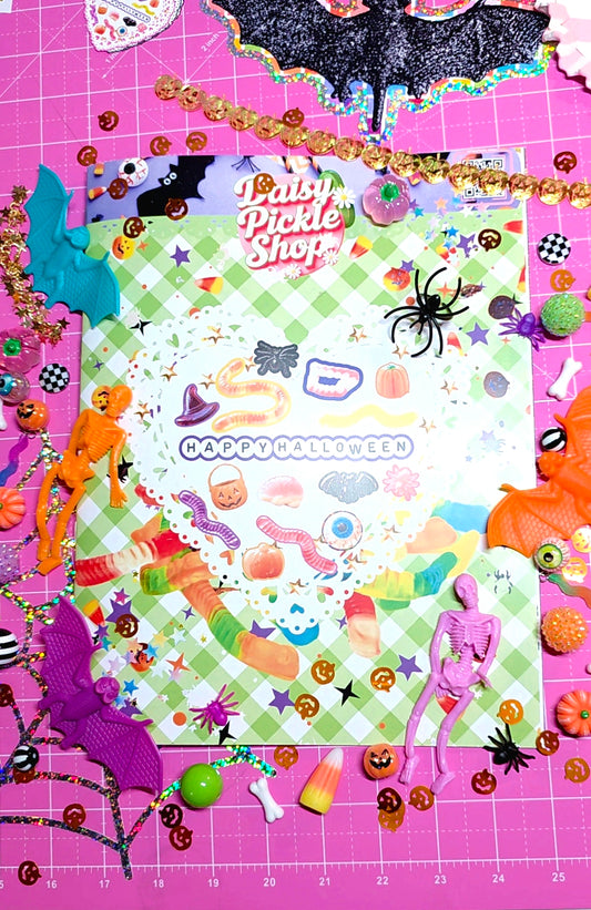 A colorful Halloween-themed scrapbook paper sheet from 'Baby Pickle Shop' surrounded by various Halloween decorations, including plastic bats, a skeleton figure, candy corn, and orange pumpkins, all arranged on a pink cutting mat.