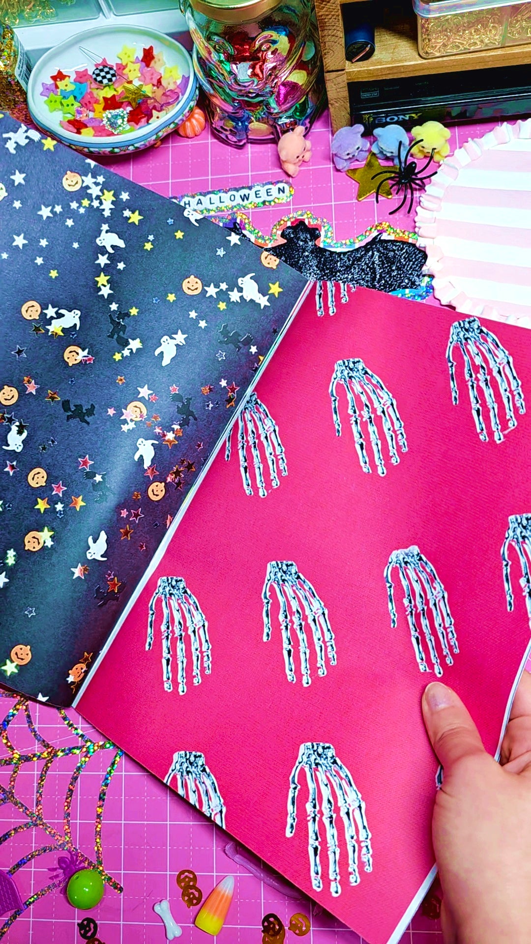 A hand holds a bright pink glittery notebook or journal page decorated with silver glitter skeleton handprints, next to another dark navy glitter page with Halloween stars and ghosts. The scene is set on a pink cutting mat surrounded by craft supplies.