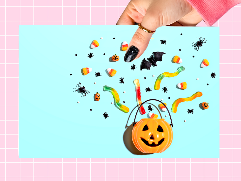 A hand with black glitter nail polish holds a square light blue card featuring a Halloween candy and spider decoration layout, set against a pink and white grid background.