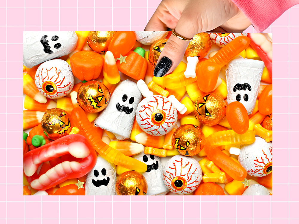 A hand with black glitter nail polish reaches in to grab a piece of orange gummy candy from a dense pile of Halloween treats, including ghosts, eyeballs, pumpkins, and corn candy, set against a pink grid background.