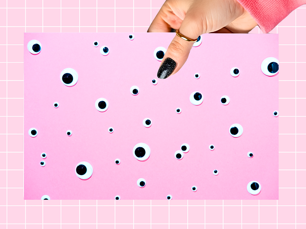 A hand with glittery black nail polish holds a pink sheet covered in scattered plastic goggle eyes, set against a pink grid background.