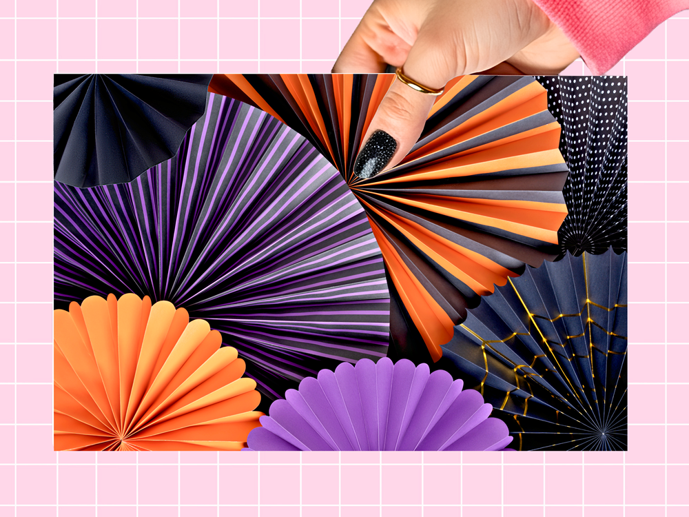 A hand with black glitter nail polish holds a rectangular sheet of colorful paper fan decorations against a pink grid background. The fans include orange, purple, black, and navy designs with striped, web, and polka-dot patterns.