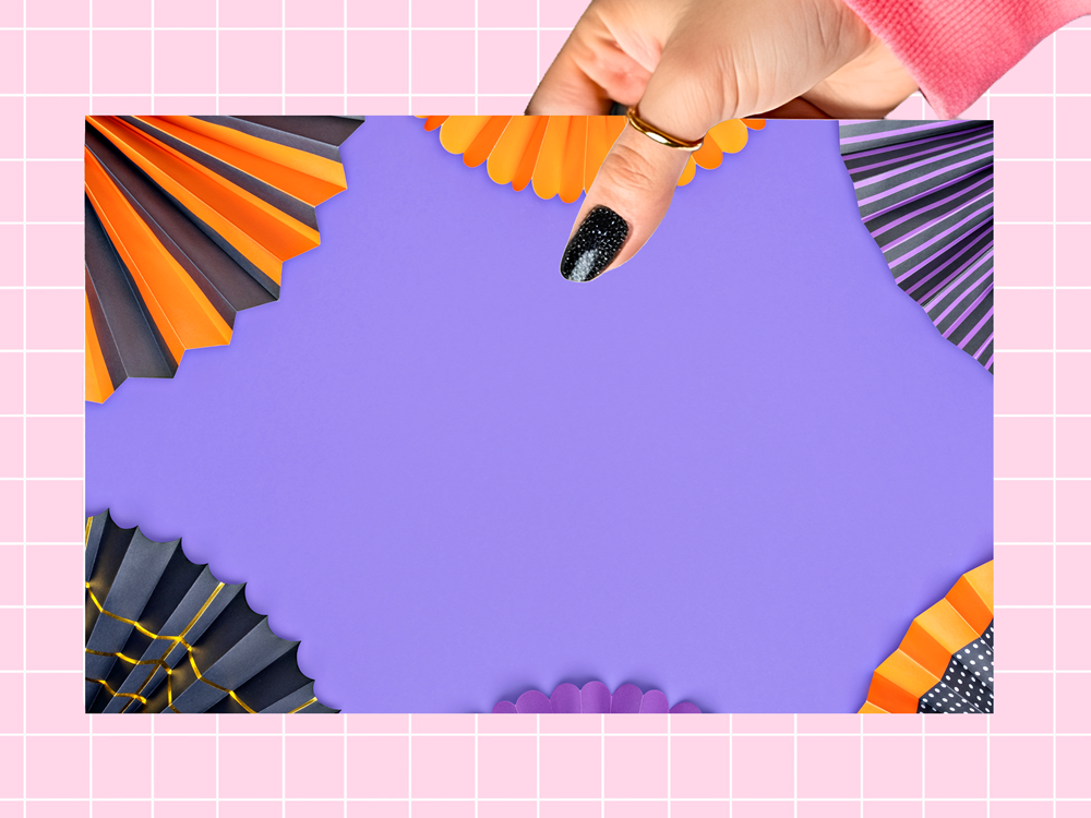 A hand with black glitter nail polish and a gold ring holds a purple rectangular card or poster with decorative paper fans in orange, black, and purple around the edges, set against a pink grid background.