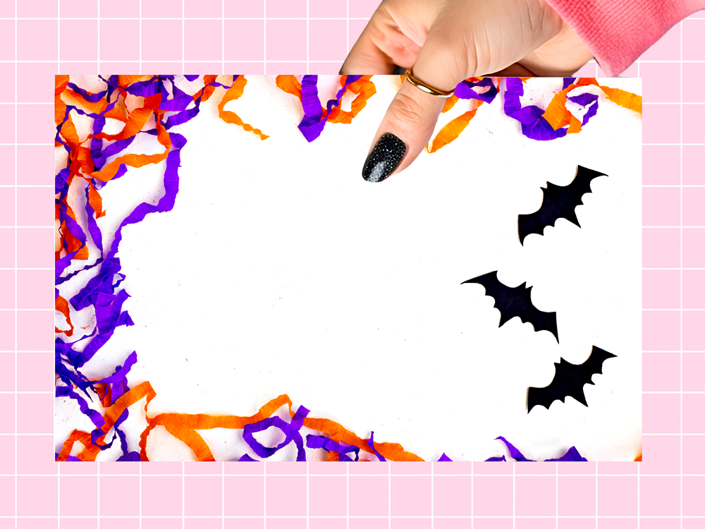 A hand with black glitter nail polish holds a white square card decorated with orange and purple shredded paper borders and three black bat silhouettes, set against a pink grid background.