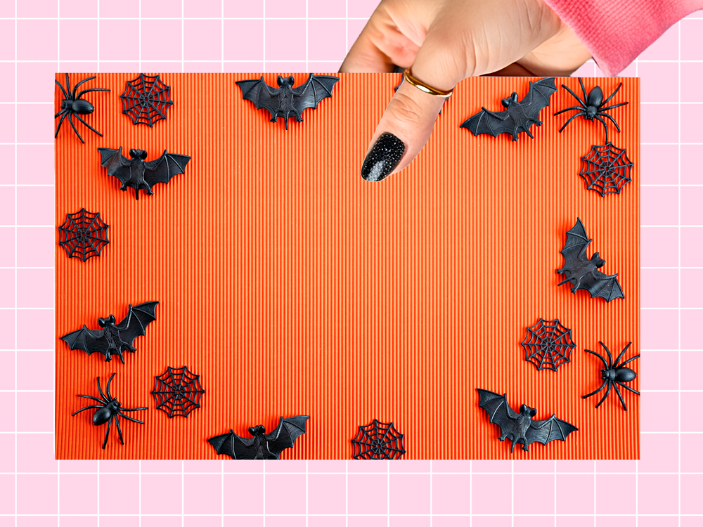 An orange ribbed rectangular mat or tray decorated with black plastic Halloween-themed items including bats, spiders, and spiderwebs, held by a hand with glittery black nail polish against a pink grid background.