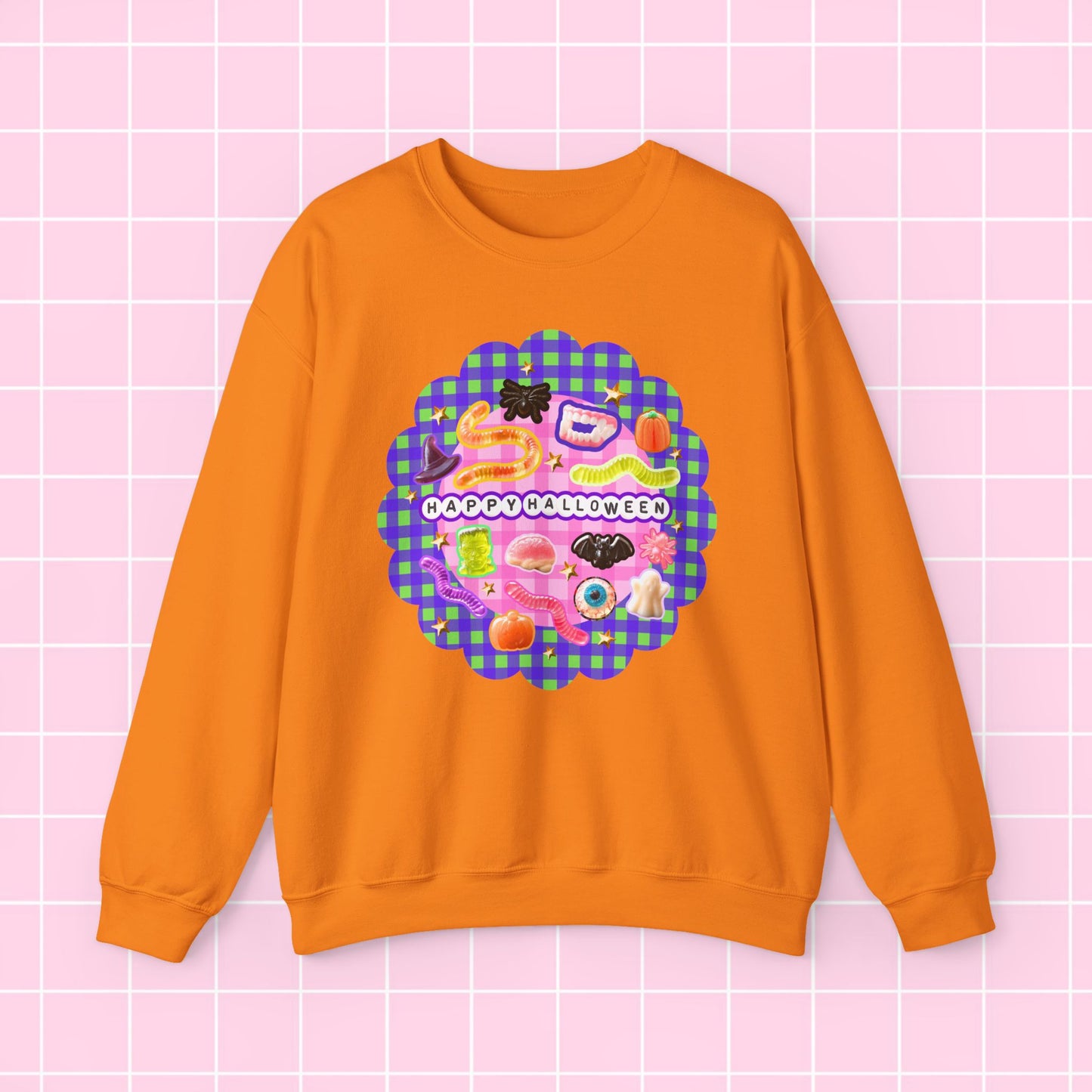 An orange crewneck sweatshirt featuring a colorful retro-style graphic with the text 'HAPPY MOTHER'S DAY' on a pink and purple gingham background, displayed against a light pink grid backdrop.