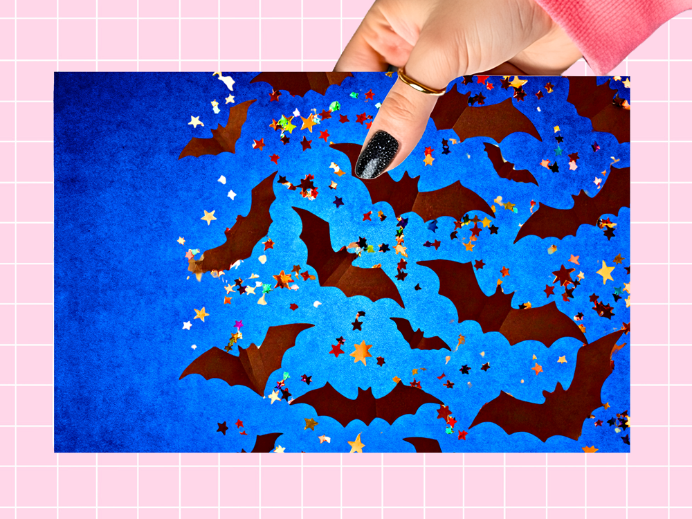 A hand with black glitter nail polish holds a rectangular blue craft sheet featuring layered brown bat silhouettes and scattered multicolored star cutouts, set against a pink grid background.