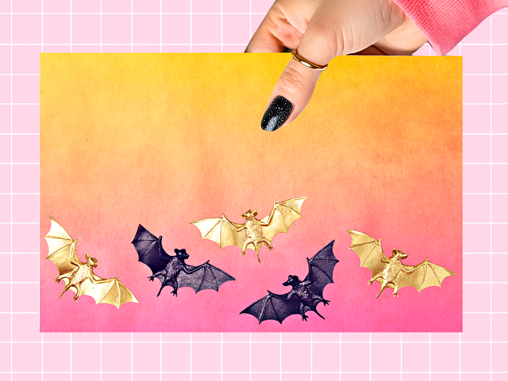 A hand with black glitter nail polish holds a square card featuring five bat illustrations in gold and black tones against a yellow-to-pink gradient background. The card is set against a pink grid backdrop.