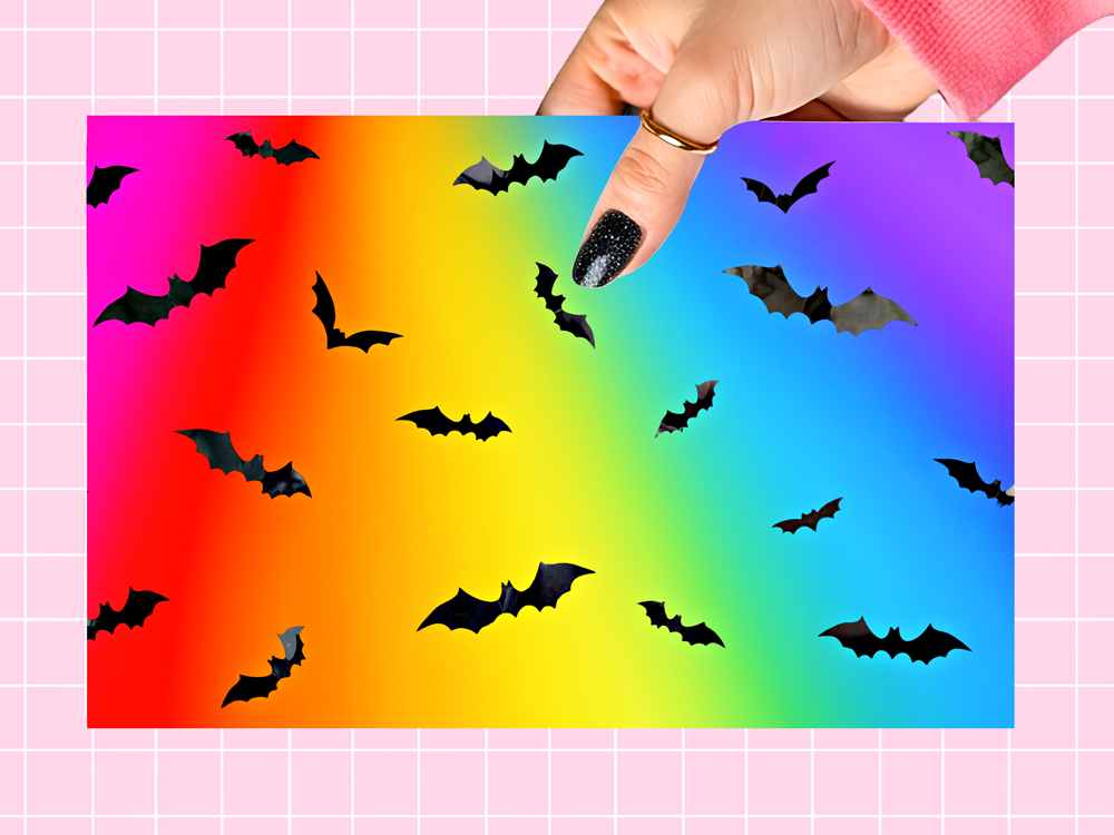 A hand with glittery black nails holds a rectangular card featuring a vibrant rainbow gradient background with scattered black bat silhouettes, set against a pink grid-patterned backdrop.