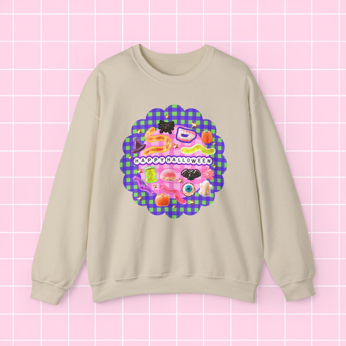A beige crewneck sweatshirt featuring a colorful retro Halloween graphic with the text 'HAPPY HALLOWEEN' on a pink and purple plaid background, displayed against a pink grid backdrop.