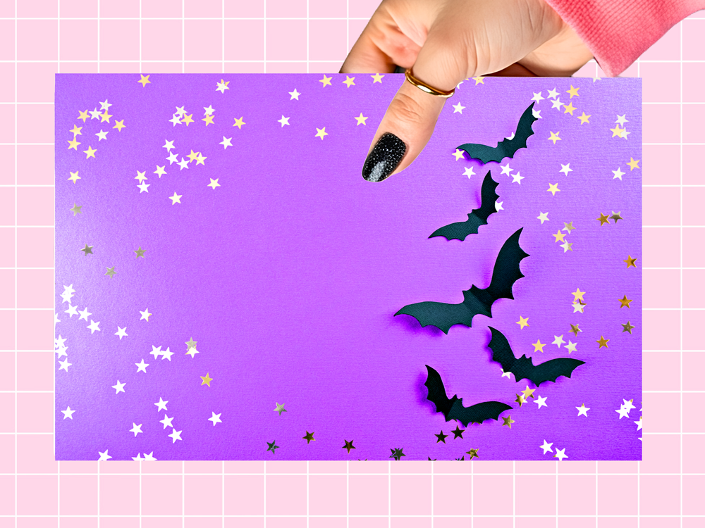 A hand with black glitter nail polish holds a purple rectangular card decorated with black bat cutouts and scattered white and gold star confetti, set against a pink grid background.