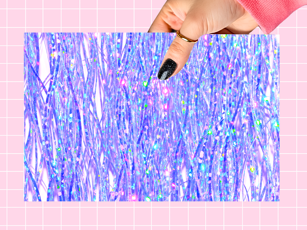 A hand with black glitter nail polish and a gold ring holds a rectangular sheet of iridescent holographic tinsel foil against a pink grid background.
