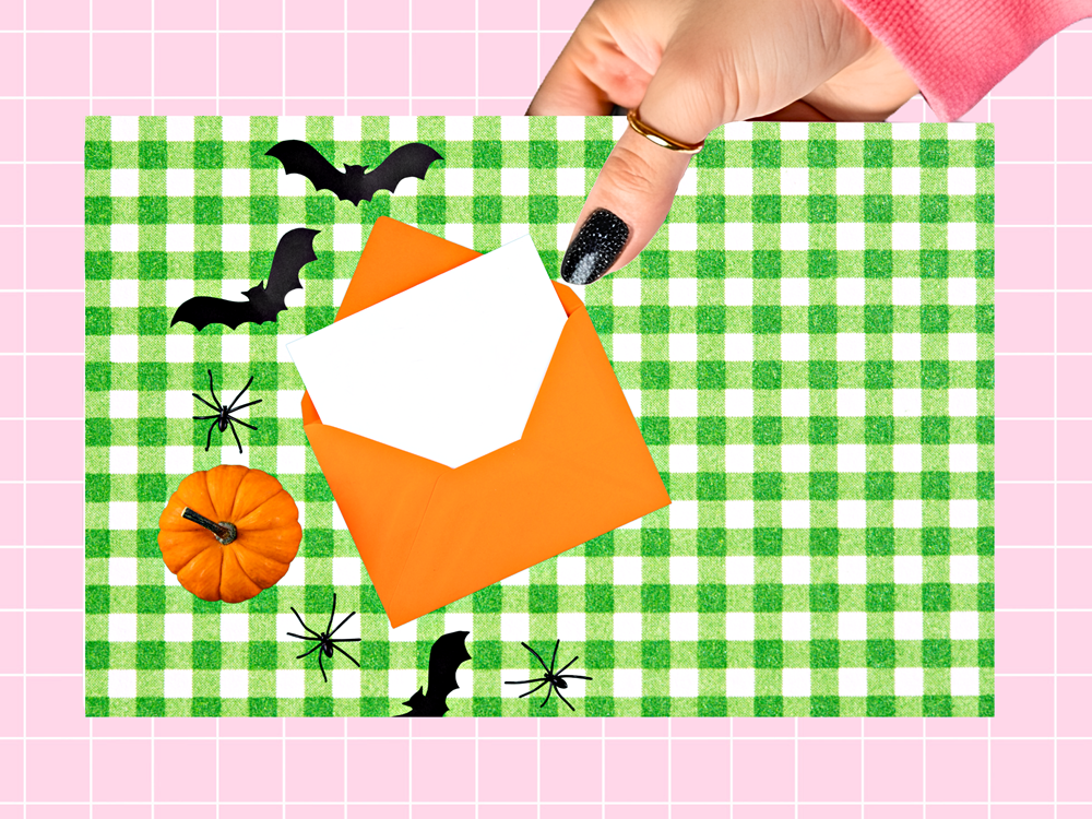 A hand with black glitter nail polish holds an orange envelope over a green and white gingham paper plate decorated with Halloween cutouts including bats, spiders, a pumpkin, and a black bat silhouette, all on a pink grid background.