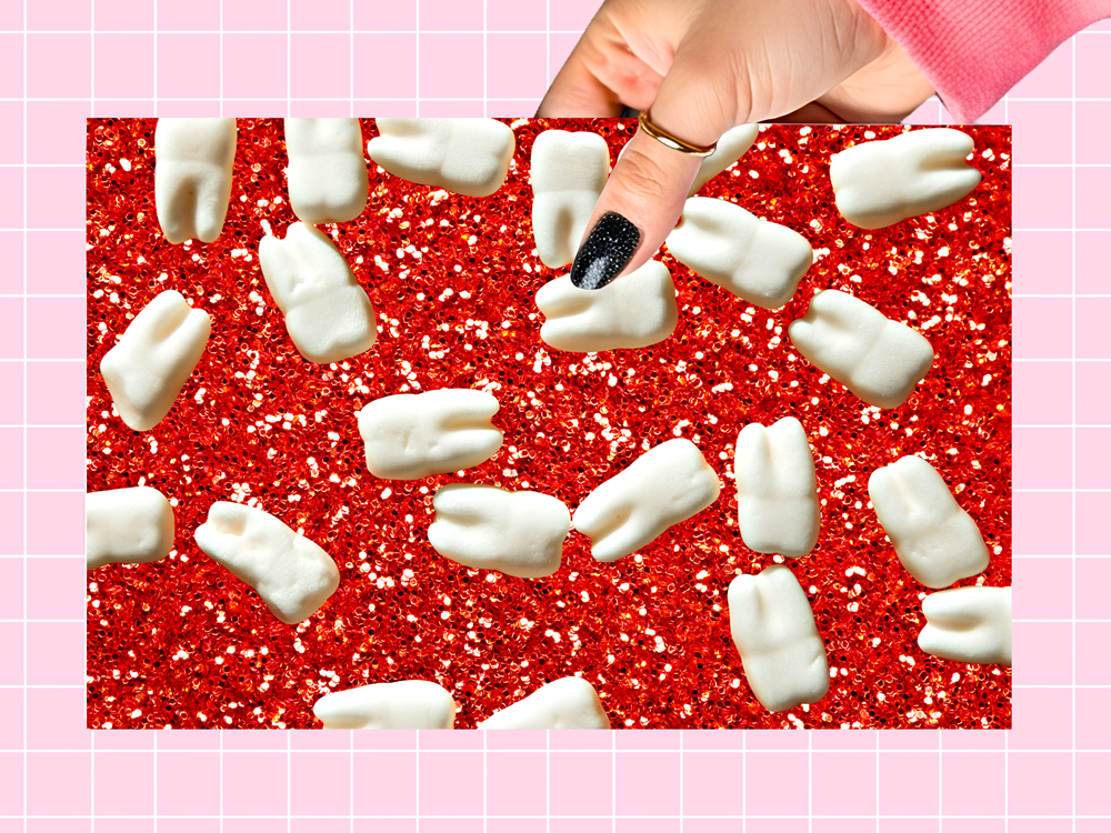 A hand with black glitter nail polish and a gold ring points toward small white tooth-shaped candies scattered on a bright red glitter background, framed by a pink grid border.
