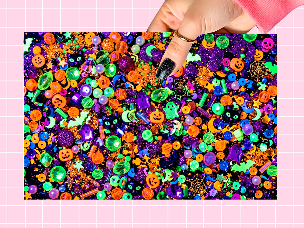 A hand with black glitter nail polish points at a colorful pile of Halloween-themed craft embellishments, including orange jack-o'-lanterns, purple ghosts, green slime beads, and multicolor glitter, set against a pink grid background.