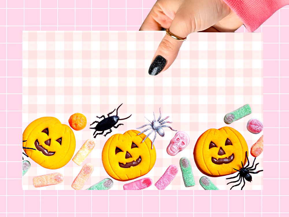 A pink and white checkered Halloween-themed cookie board featuring three jack-o'-lantern cookies, candy corn, gummy candies, plastic spiders, and a skull, held by a hand with black glitter nail polish against a pink grid background.