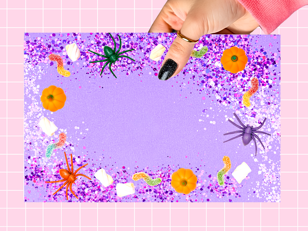 A hand with black glitter nail polish holds a square purple Halloween craft card decorated with glitter, plastic spiders, worms, and pumpkins, set against a pink grid background.