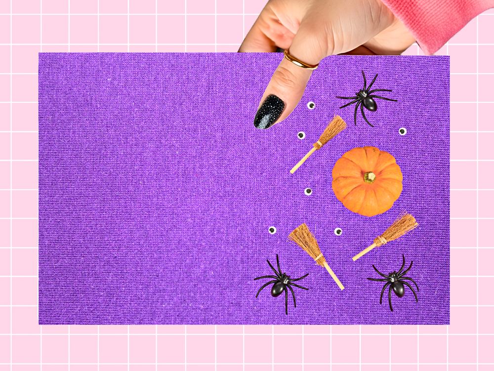 A hand with black glitter nail polish holds a purple textured square card featuring Halloween-themed illustrations of black spiders, orange pumpkins, and small broomsticks, set against a pink grid background.