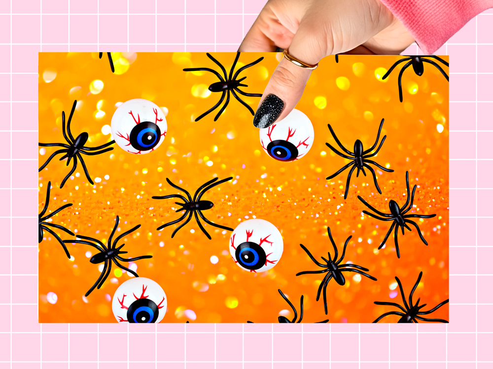 A hand wearing a pink sleeve and black glitter nail polish points to a Halloween craft sheet filled with black plastic spiders and white eyeballs on a glittery orange background, set against a pink grid backdrop.