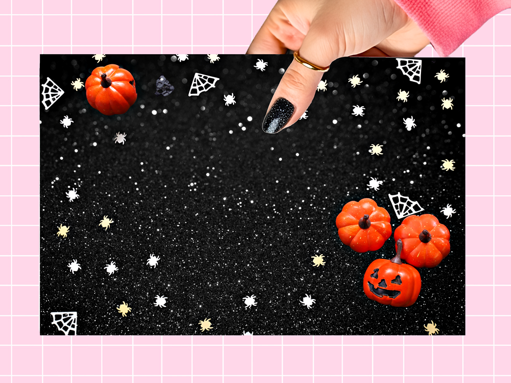 A rectangular black glittery card or backdrop with Halloween-themed graphics, held by a hand. The design features small orange jack-o'-lantern pumpkins, white spiderwebs, and white/gold stars on a black glittery background.