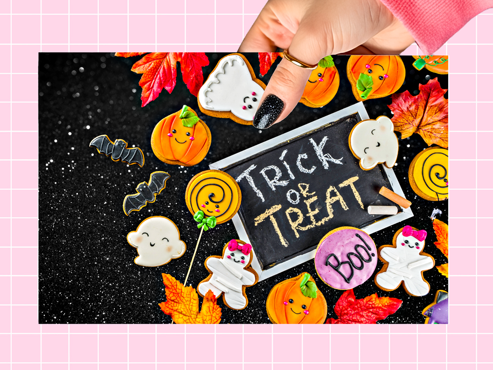 A hand with black glitter nail polish holds a rectangular blackboard-style sign that reads “Trick or Treat,” surrounded by Halloween-themed decorated cookies including ghosts, pumpkins, bats, and a purple Boo cookie. The scene is framed by orange and red autumn leaves on a black glitter background, with a pink grid border around the image.