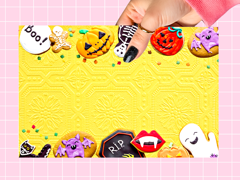 A hand with black glitter nail polish holds a square yellow Halloween-themed cookie cutter template or mat against a pink grid background. The yellow surface features embossed geometric patterns and is decorated with various colorful Halloween cookies, including pumpkins, ghosts, bats, a skeleton, vampire lips, and a 'Boo!' speech bubble.