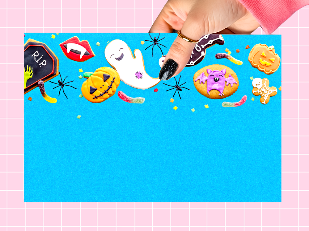 A hand with black glitter nail polish holds a blue card decorated with Halloween-themed cookies and illustrations, set against a pink grid background.