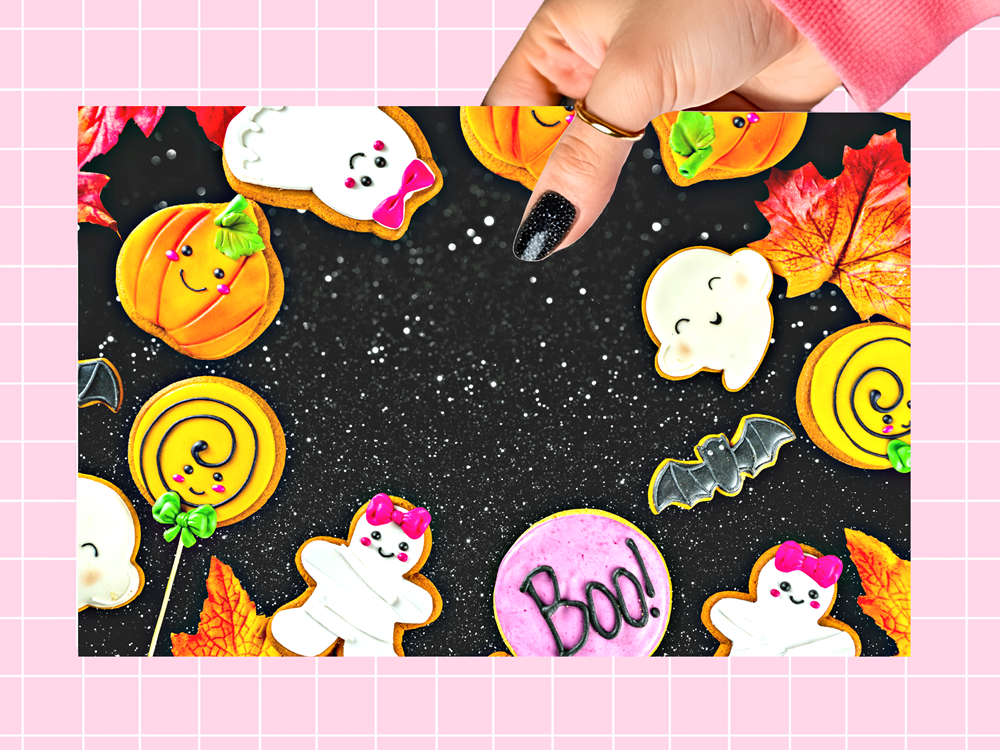 A hand with black glitter nail polish holds a square Halloween-themed cookie board featuring decorated sugar cookies shaped like pumpkins, ghosts, lollipops, bats, and a pink 'Boo!' cookie on a black background with white speckles. The photo is framed by a pink grid border.