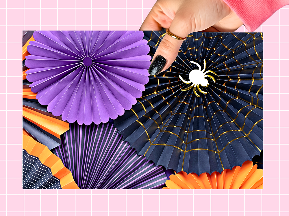 A hand with black glitter nail polish holds a rectangular sheet of layered paper fan decorations. The set includes purple, navy blue with gold web patterns, orange, and dark purple striped fans, decorated with a white spider cutout.