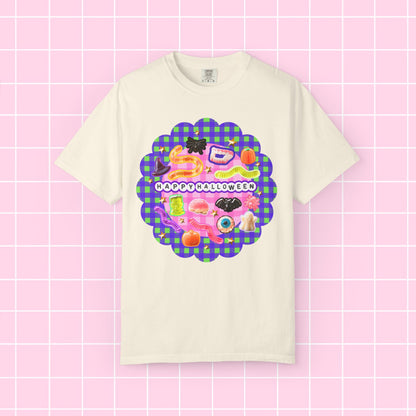 A cream-colored short-sleeve t-shirt with a colorful 'Happy Halloween' graphic on the front, displayed against a pink grid background.