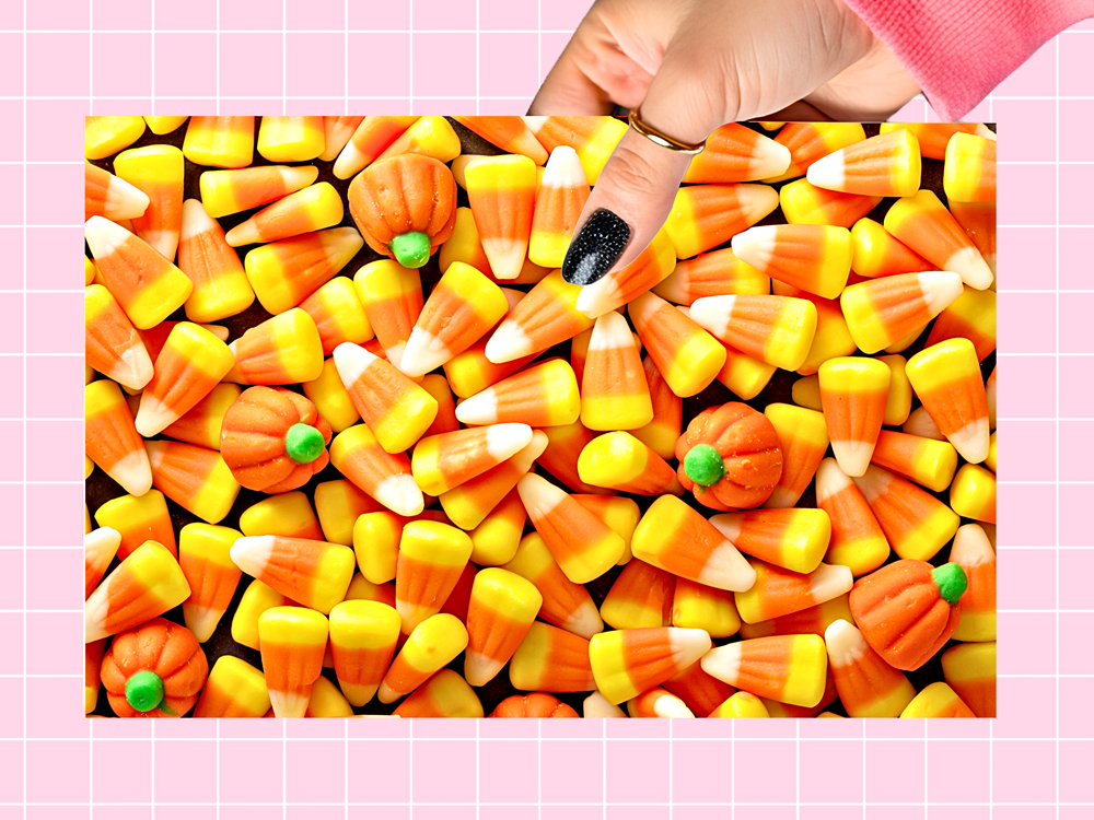 A close-up of a hand with black glitter nail polish reaching into a pile of candy corn candies, with small orange pumpkin-shaped candies and green tops mixed in. The scene is framed by a pink grid background.