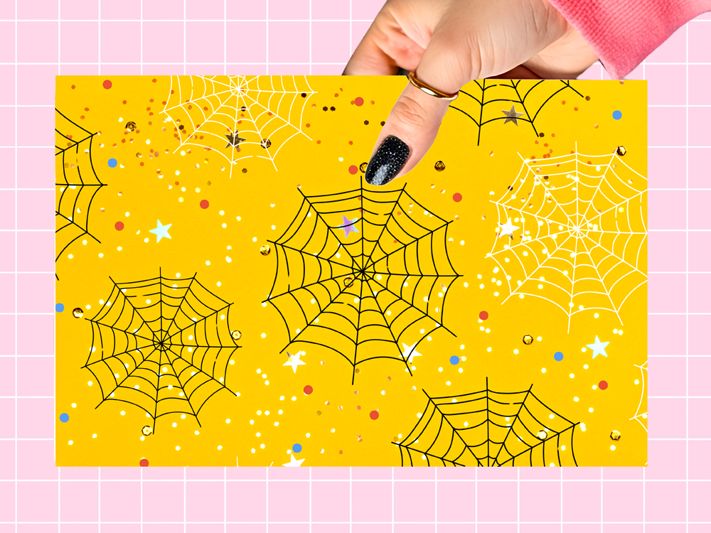 A hand with black glitter nail polish holds a square yellow card featuring a Halloween spiderweb pattern with black spiderwebs, white stars, and small colorful glitter dots. The background is a pink grid pattern.