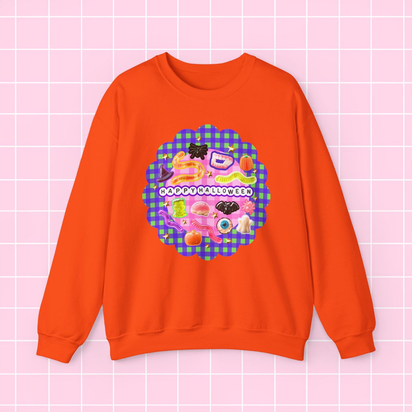 An orange crewneck sweatshirt featuring a colorful retro-style graphic with the text 'HAPPY HOLIDAYS' on a pink and purple gingham background, displayed on a light pink grid backdrop.