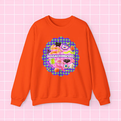 An orange crewneck sweatshirt featuring a colorful retro-style graphic with the text 'HAPPY HOLIDAYS' on a pink and purple gingham background, displayed on a light pink grid backdrop.