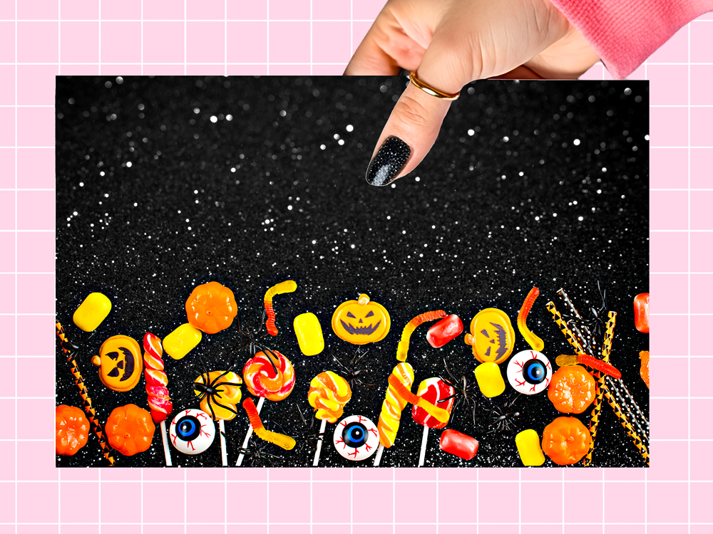 A rectangular black backdrop printed with a Halloween candy border, held up by a hand with glittery black nail polish against a pink grid background.