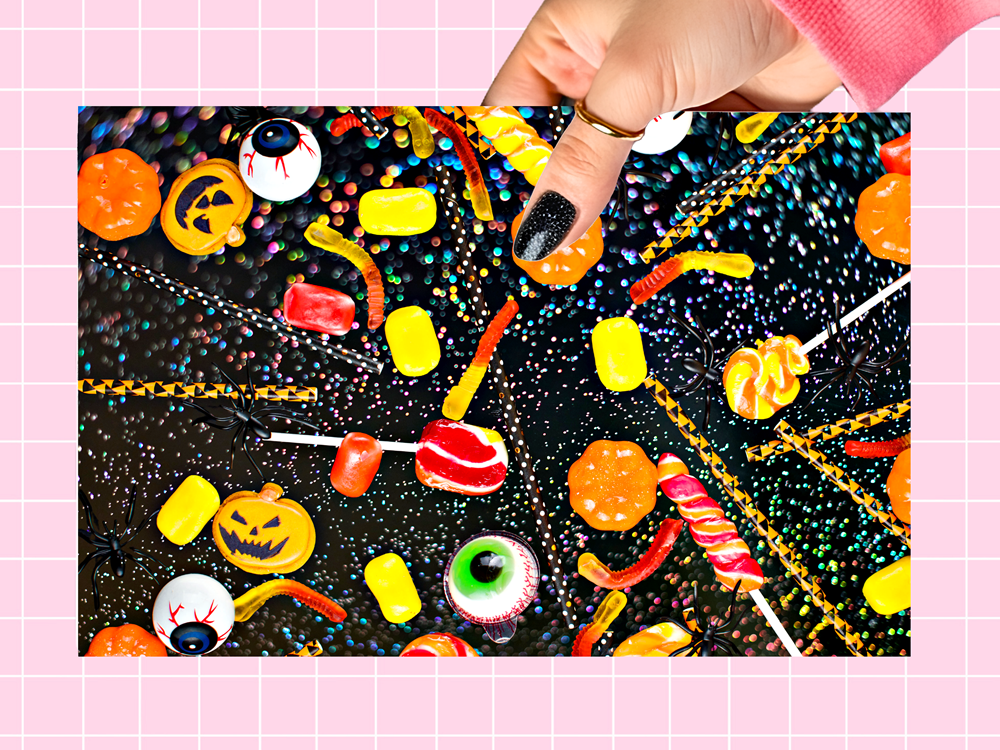 A hand with black glitter nail polish holds a rectangular card featuring a colorful Halloween candy illustration on a black glittery background, set against a pink grid backdrop.