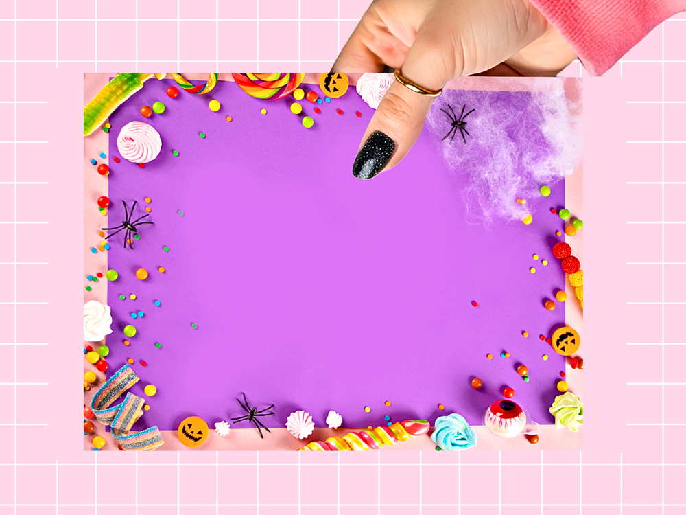A hand with black glitter nail polish holds a purple rectangular card or poster decorated with colorful Halloween-themed candy, sprinkles, spiderwebs, and smiley faces on a pink grid background.