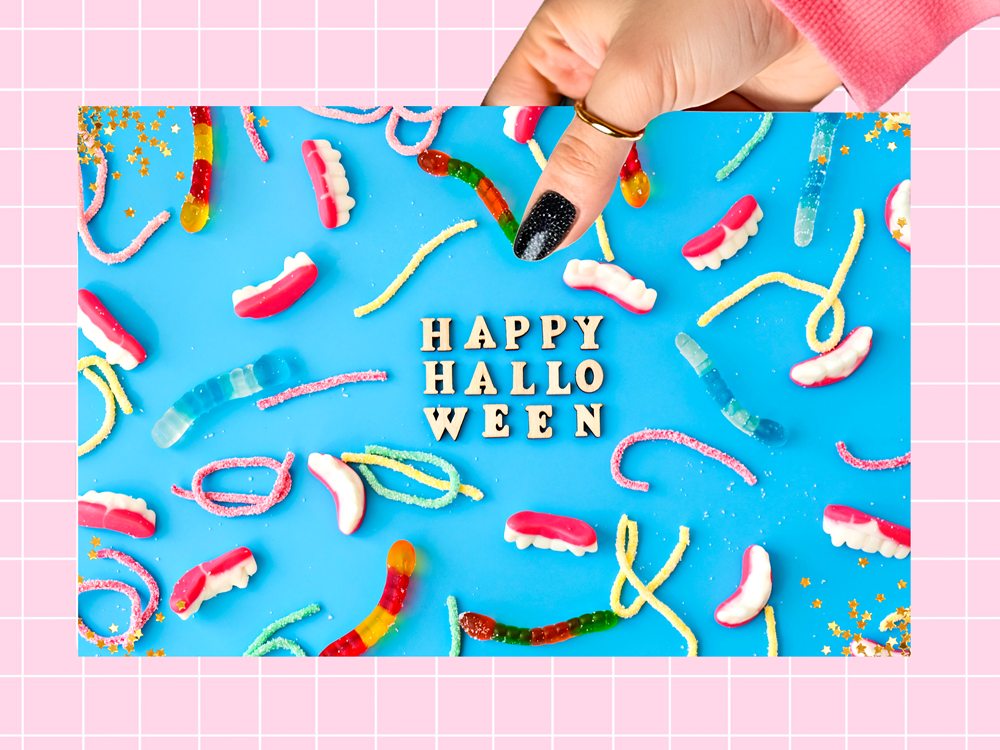 A hand with black glitter nail polish holds a pink card featuring colorful Halloween-themed gummy candies and the words 'HAPPY HALLOWEEN' in white letters, set against a bright blue background with a pink grid border.