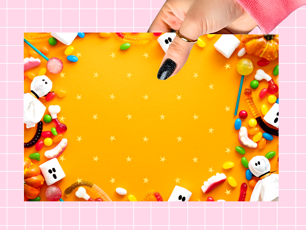 A hand with black glitter nail polish holds a square orange card decorated with Halloween-themed candies, gummy worms, marshmallows, and ghost candies, set against a pink grid background.