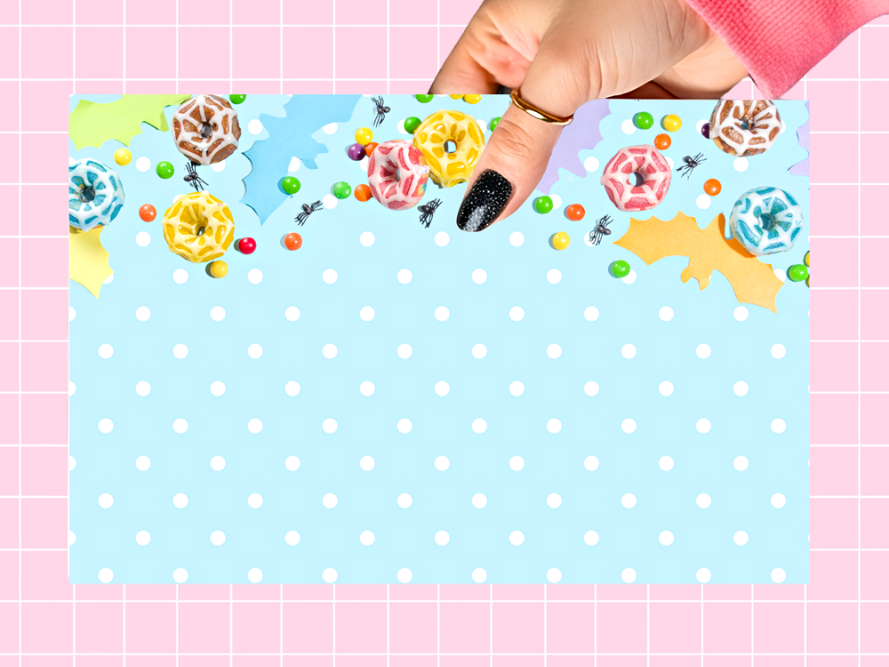 A hand with black glitter nail polish holds a rectangular light blue card with white polka dots and colorful candy-themed illustrations along the top, set against a pink grid background.
