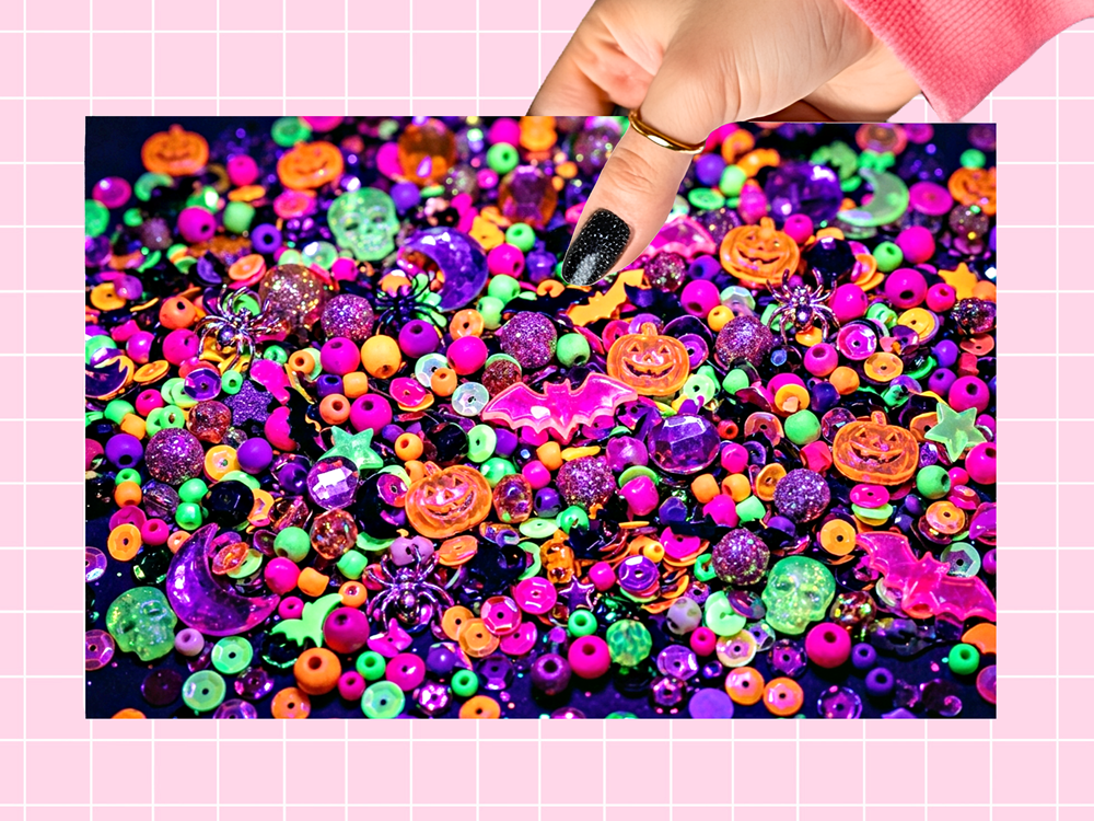 A close-up of a hand with black glitter nail polish touching a pile of colorful Halloween-themed craft beads, sequins, and decorative embellishments. The beads feature jack-o'-lanterns, spiders, pumpkins, stars, and glitter finishes in vibrant neon colors.