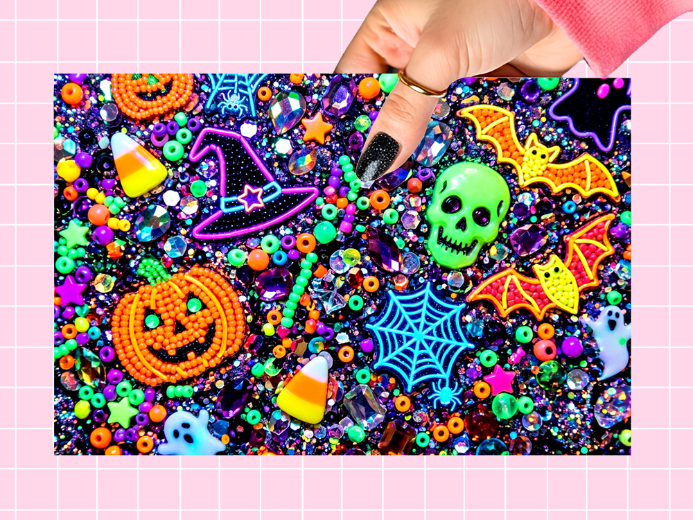 A hand with black glitter nail polish holds a square sheet of colorful Halloween-themed craft beads and embellishments. The sheet features orange beaded pumpkins, yellow and orange beaded bats, a purple beaded witch hat, a green beaded skull, candy corn, spiderwebs, ghosts, and a black glitter background.